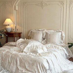 May include: A cream-colored bedding set featuring a duvet cover and matching pillow shams with ruffled edges and intricate lace-like floral embroidery. The fabric has a smooth finish, and the set includes multiple pillows with layered ruffles.