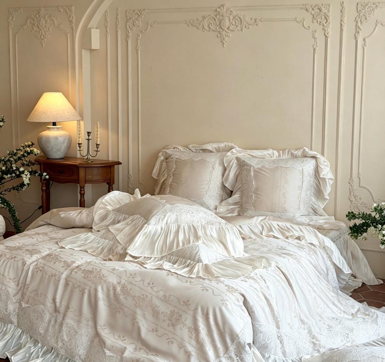 May include: A cream-colored bedding set featuring a duvet cover and matching pillow shams with ruffled edges and intricate lace-like floral embroidery. The fabric has a smooth finish, and the set includes multiple pillows with layered ruffles.