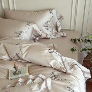 May include: A beige bedding set featuring a duvet cover and two pillowcases with ruffled edges and delicate floral embroidery. A small silver candlestick with a taper candle and an open book rest on the smooth, satin-like fabric surface.