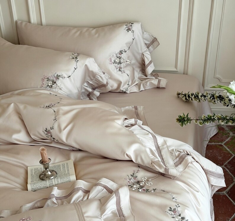 May include: A beige bedding set featuring a duvet cover and two pillowcases with ruffled edges and delicate floral embroidery. A small silver candlestick with a taper candle and an open book rest on the smooth, satin-like fabric surface.