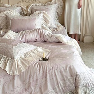 May include: A bedding set in soft mauve featuring a delicate white lace pattern and ruffled cream-colored fabric borders. The set includes multiple pillows with ruffled edges and a matching duvet cover, all arranged on a bed with a small candle.