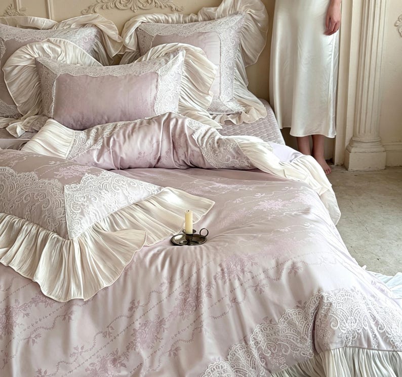 May include: A bedding set in soft mauve featuring a delicate white lace pattern and ruffled cream-colored fabric borders. The set includes multiple pillows with ruffled edges and a matching duvet cover, all arranged on a bed with a small candle.