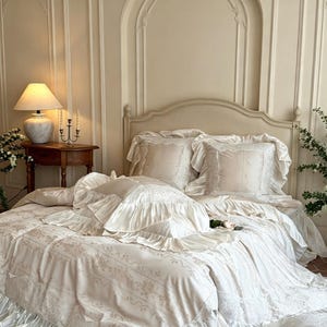 May include: A cream-colored bedding set featuring a duvet cover and matching pillow shams with ruffled edges and intricate lace trim. The fabric displays a subtle floral pattern, and the set includes multiple pillows with ruffled borders.