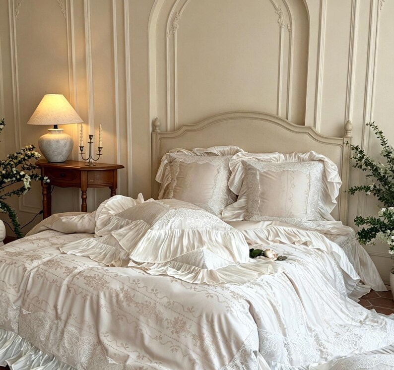 May include: A cream-colored bedding set featuring a duvet cover and matching pillow shams with ruffled edges and intricate lace trim. The fabric displays a subtle floral pattern, and the set includes multiple pillows with ruffled borders.