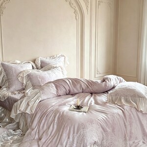 May include: A light lavender bedding set featuring a duvet cover and pillowcases with intricate white lace-like patterns and ruffled edges. A small candle in a metal holder sits on a book placed in the center of the bedspread.