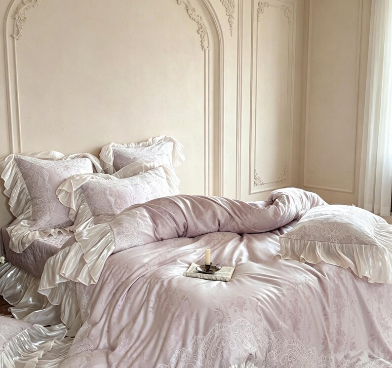 May include: A light lavender bedding set featuring a duvet cover and pillowcases with intricate white lace-like patterns and ruffled edges. A small candle in a metal holder sits on a book placed in the center of the bedspread.