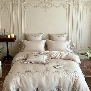 May include: A beige bedding set featuring a duvet cover, two standard pillowcases, and one small rectangular accent pillow. The fabric is adorned with delicate floral embroidery along the edges and ruffled trim on the pillowcases and duvet.