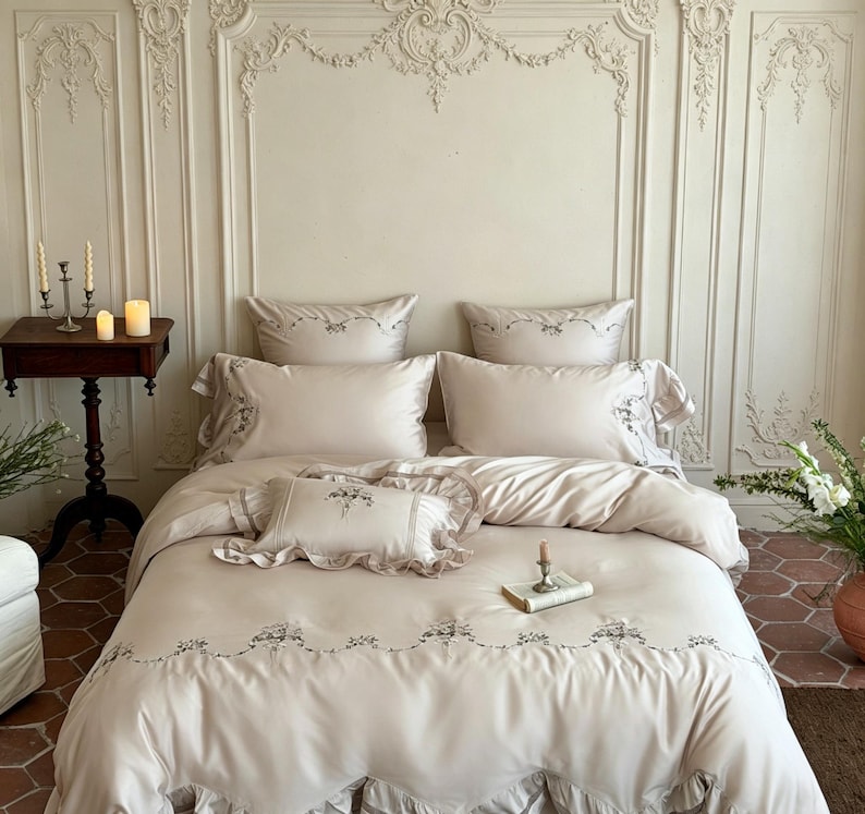 May include: A beige bedding set featuring a duvet cover, two standard pillowcases, and one small rectangular accent pillow. The fabric is adorned with delicate floral embroidery along the edges and ruffled trim on the pillowcases and duvet.