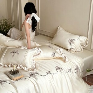 May include: A cream-colored bedding set featuring a duvet cover and matching pillowcases with delicate floral embroidery and ruffled edges. The fabric has a smooth, satin-like finish, and the design includes intricate botanical motifs.