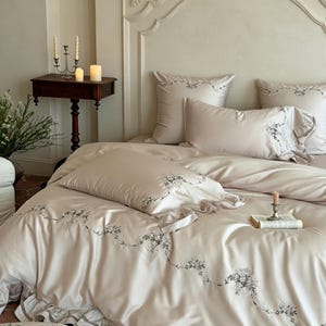 May include: A beige bedding set featuring a duvet cover and pillowcases with delicate grey floral embroidery and ruffled edges. The fabric has a smooth, satin-like finish, and the set includes matching square and rectangular pillows.