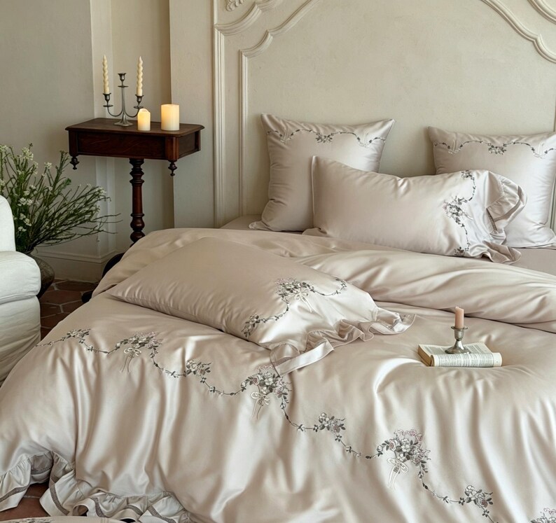 May include: A beige bedding set featuring a duvet cover and pillowcases with delicate grey floral embroidery and ruffled edges. The fabric has a smooth, satin-like finish, and the set includes matching square and rectangular pillows.