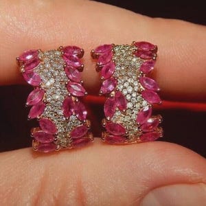 Könnte beinhalten: Ein Paar goldfarbene Ohrringe. Jeder Ohrring hat einen Mittelteil aus kleinen, klaren Steinen, umgeben von leuchtend rosa, marquiseförmigen Edelsteinen. Die Ohrringe sind vor einem roten Hintergrund platziert.
