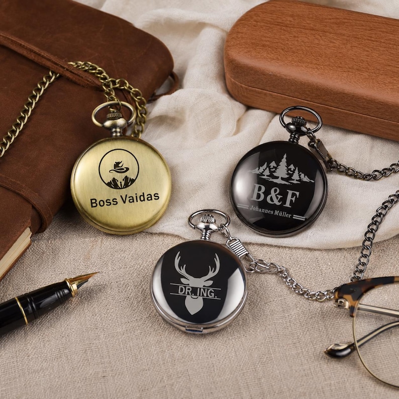 May include: Three round pocket watches with attached chains. One is brushed gold with a mountain logo and the text Boss Vaidas. One is black with a forest graphic and the text B&F Johannes M&uuml;ller. One is silver with a deer head and DR. ING. text.