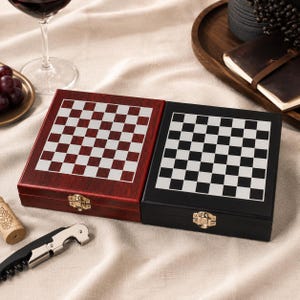 Puede incluir: Dos juegos de ajedrez de madera, uno en acabado rojo oscuro y otro en negro, cada uno con un patrón de cuadros plateado y blanco en la tapa. Los juegos se cierran con un cierre dorado. Un sacacorchos y un corcho están en primer plano.
