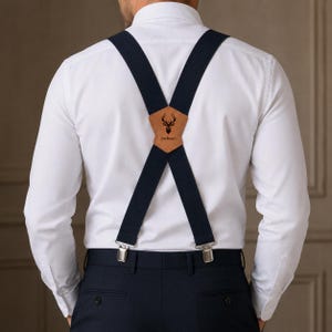 Peut inclure: Bretelles bleu marine foncé avec une pièce dorsale en cuir beige ornée d'une silhouette de tête de cerf noire et du nom Jackson imprimé en dessous. Les bretelles sont portées sur une chemise blanche et un pantalon bleu marine foncé.