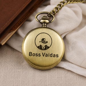 May include: A round, antique brass-toned pocket watch with a matching metal chain. The front cover features a circular graphic of a stylized animal silhouette above mountain peaks, with the text Boss Vaidas printed clearly below the graphic.