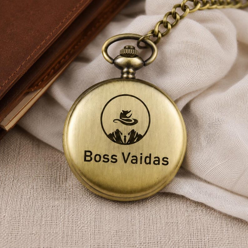 May include: A round, antique brass-toned pocket watch with a matching metal chain. The front cover features a circular graphic of a stylized animal silhouette above mountain peaks, with the text Boss Vaidas printed clearly below the graphic.
