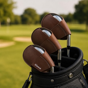 Könnte beinhalten: Drei braune Golfschlägerhauben mit Steppnähten, die jeweils einen silbernen Metallschlägerkopf umschließen. Die vorderste Haube zeigt das Wort Name in weißer Schreibschrift. Die Hauben befinden sich in einer schwarzen Golftasche mit silbernen Beschlägen.