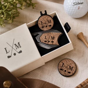 Könnte beinhalten: Ein Golf-Geschenkset mit einer weißen Box mit den Initialen "L M" und Golfschläger-Grafiken. Darin befinden sich ein Pitchgabel-Werkzeug und ein Ballmarker mit denselben Initialen. Ein Golfball und ein Holz-Tee sind ebenfalls vorhanden.