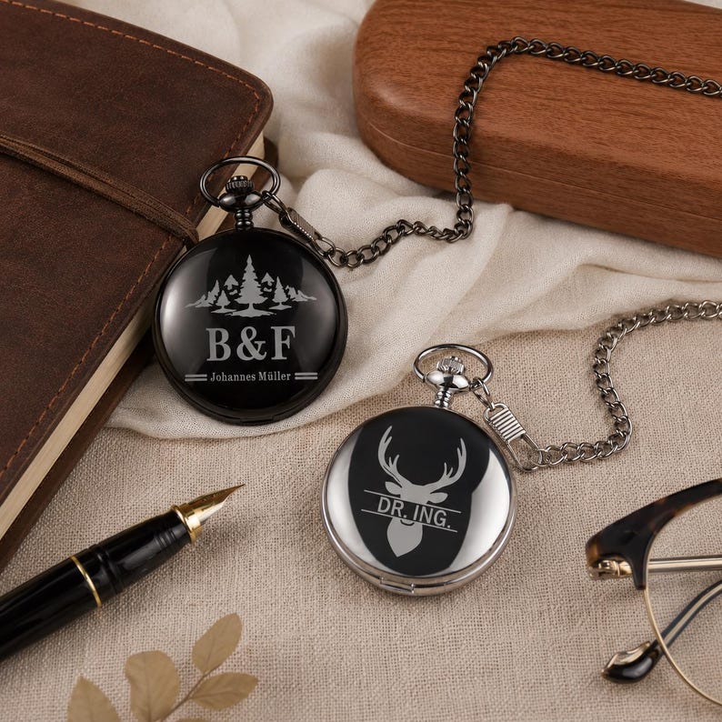 May include: Two round pocket watches with attached metal chains. One watch is black with a white engraving of pine trees, the letters B&F, and the text Johannes M&uuml;ller. The other watch is silver with a white engraving of a deer head and the text DR. ING.