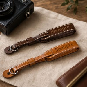 Peut inclure: Deux lanières d'appareil photo en cuir avec un design en boucle, des coutures blanches et un anneau en métal argenté à une extrémité. Chaque lanière porte le mot "Siempre" gravé sur la partie large. L'une est marron foncé, l'autre est beige.