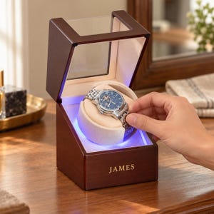 May include: A dark brown wooden watch winder box with a clear glass top and a beige interior cushion. The front of the box features the name JAMES engraved in gold lettering. A silver-tone analogue watch with a blue face is mounted inside.