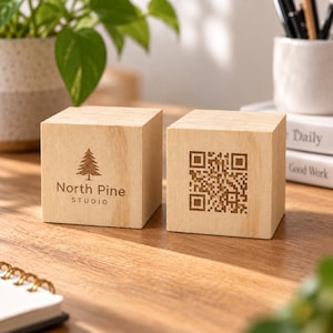 May include: Two light-colored wooden cubes featuring laser-engraved designs. The left cube displays a pine tree icon above the text North Pine Studio. The right cube features a square QR code pattern. Both cubes have a smooth, natural wood grain.