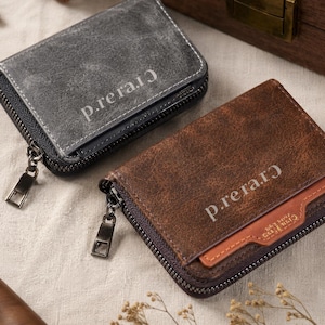 May include: Two leather wallets, one grey and one brown, with a zip closure. The grey wallet has the text "p.rerarɔ" printed on it. The brown wallet has the same text and a small card slot. Both wallets are on a neutral-coloured surface.