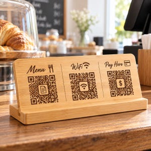 May include: A rectangular bamboo sign featuring three engraved sections with QR codes. The left section is labelled Menu with a fork and knife icon. The centre section is labelled WiFi with a signal icon. The right section is labelled Pay Here with a card icon.