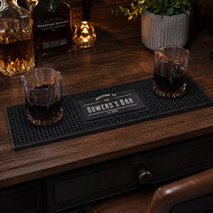 Puede incluir: Una alfombrilla de bar rectangular de goma negra con una superficie texturizada de tacos y una placa central rectangular con el texto impreso en blanco "WELCOME TO THE BOWERS'S BAR est. 2026". La alfombrilla está sobre una superficie de madera oscura.