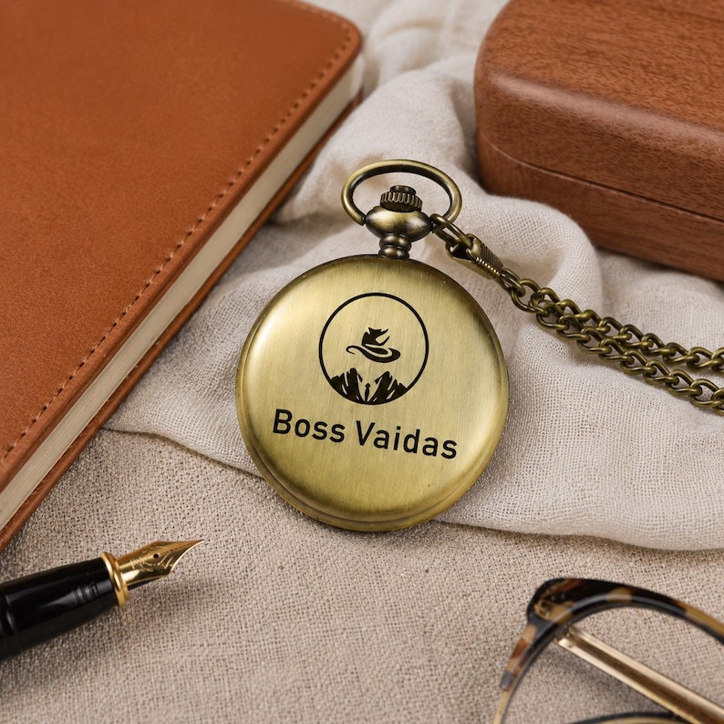 May include: A round, antique brass-toned pocket watch with a metal chain. The front cover features a circular graphic of a mountain range and a stylized hat, with the text "Boss Vaidas" printed in black lettering below the circular design.