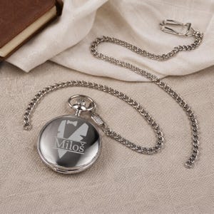 May include: A polished silver-tone pocket watch with a matching metal chain and a clip. The front of the watch features an engraved graphic of a tuxedo jacket with a bow tie and the name Milo&scaron; printed in a clear, sans-serif font.