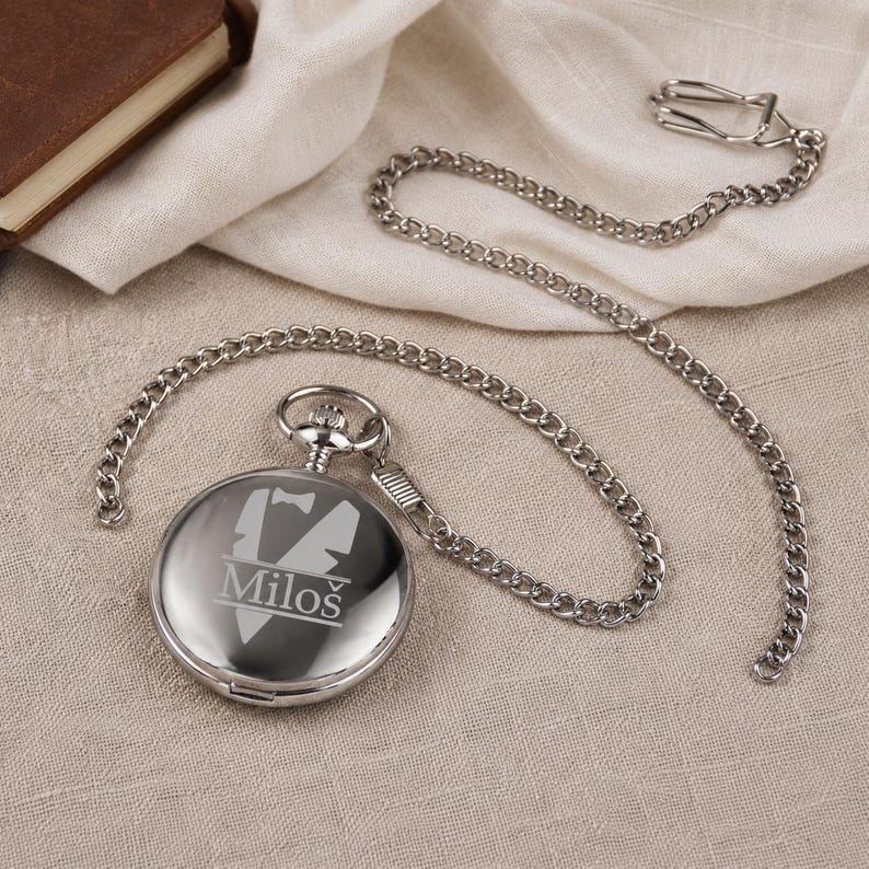 May include: A polished silver-tone pocket watch with a matching metal chain and a clip. The front of the watch features an engraved graphic of a tuxedo jacket with a bow tie and the name Milo&scaron; printed in a clear, sans-serif font.