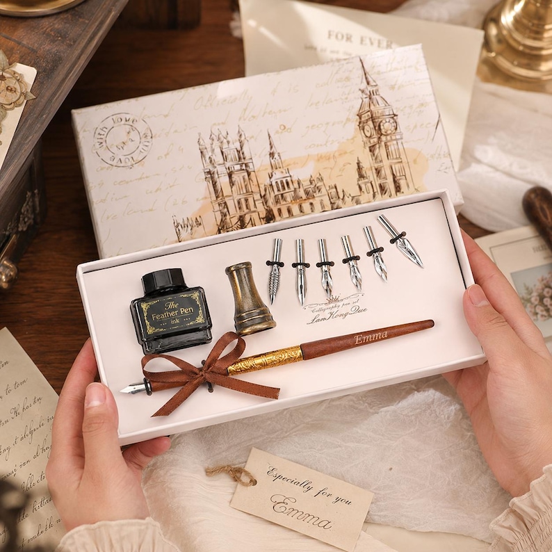 Peut inclure: Un ensemble de calligraphie dans une bo&icirc;te blanche comprenant un stylo en bois marron avec une poign&eacute;e dor&eacute;e, un flacon d'encre noire &eacute;tiquet&eacute; The Feather Pen, un porte-plume en laiton et cinq plumes argent&eacute;es. Le couvercle porte le texte LamHongDuo.