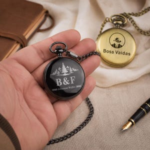 May include: A black pocket watch with a chain featuring a white engraved forest graphic, the text B&F, and the name Johannes M&uuml;ller. A second brass-colored pocket watch with a chain displays a logo and the text Boss Vaidas.