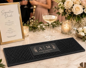 Puede incluir: Una alfombrilla de bar rectangular de goma negra con un panel central negro que presenta un monograma blanco de las letras A y M, rodeado de ilustraciones florales y el texto Alexa & Michael 06.22.2024, con una superficie texturizada de puntos.