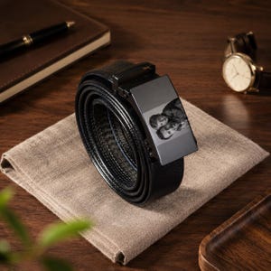 Op de afbeelding: Een zwarte leren riem met een rechthoekige metalen gesp voorzien van een aangepaste, monochroom gegraveerde foto van twee personen. De riem heeft een gestructureerd oppervlak en een gladde, donkere afwerking.