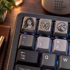 Op de afbeelding: Een zwart mechanisch toetsenbord met drie aangepaste metalen toetsen met lasergravures: een portret van een persoon, een golden retriever en een berglandschap met een zon. Het toetsenbord heeft grijze en zwarte toetsen met blauwe achtergrondverlichting.