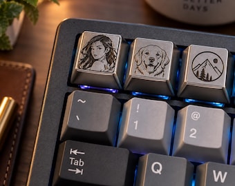 May include: A black mechanical keyboard featuring three custom metallic keycaps with laser-engraved designs: a portrait of a person, a golden retriever dog, and a mountain landscape with a sun. The keyboard has grey and black keys with blue backlighting.