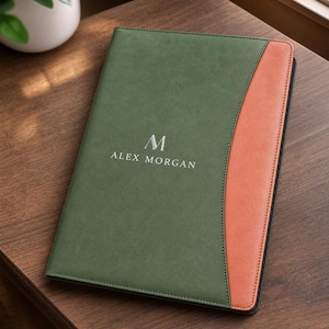 May include: A rectangular notebook with a matte olive green cover featuring a curved terracotta orange side panel. The centre of the cover displays the letter M above the text ALEX MORGAN in a clean, white, sans-serif font with visible stitching.