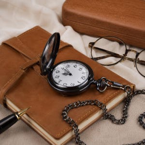 May include: A black pocket watch with an open lid, a white face, and black numerals, attached to a matching black metal chain. The watch rests on a tan leather-bound journal, accompanied by a fountain pen and round-rimmed glasses.
