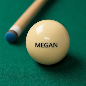 May include: A smooth, cream-colored billiard cue ball featuring the name MEGAN printed in black capital letters, positioned next to a wooden pool cue with a blue tip, resting on a green felt surface.
