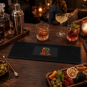 May include: A rectangular black rubber bar mat with a textured surface and a central logo featuring a house icon, two glasses, and the text HOME VIBE HOUSE PARTY. The mat is positioned on a dark wooden surface among various cocktail glassware.