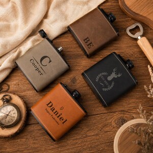 Peut inclure: Quatre flasques personnalisées en cuir marron, beige et noir. L'une porte l'initiale "C" et le nom "Cooper". Une autre porte le nom "Daniel" et les mots "Super Padrinho". Les deux autres flasques ont les initiales "BE" et un motif de tête de cerf.