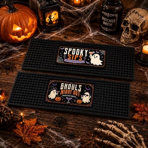 May include: Two rectangular black rubber bar mats with raised nubs. The top mat features a white ghost holding a cocktail with the text "SPOOKY SIPS Cheers Boos and Brews". The bottom mat shows two ghosts and pumpkins with the text "GHOULS NIGHT OUT Raise Your Spirits".