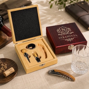 Puede incluir: Un juego de herramientas para vino de madera con sacacorchos, tapón y vertedor. El juego está en una caja de madera de color claro con un interior negro. También se ve una caja roja oscura separada con el nombre "STRAHINJA".