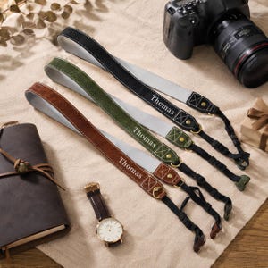 May include: Three camera straps in black, olive green, and brown, each featuring a light grey underside, gold-tone metal hardware, and the name Thomas embossed in white. The straps include black adjustable nylon webbing and plastic connectors.