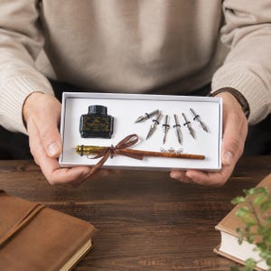May include: A white rectangular gift box containing a black ink bottle labelled The Feather Pen, five silver metal calligraphy nibs, and a brown wooden pen with a gold-coloured tip, tied with a brown ribbon and engraved with the name Gabriel.