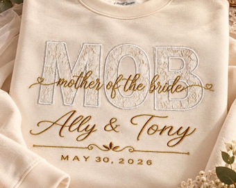 Könnte beinhalten: Ein cremefarbenes Sweatshirt mit Rundhalsausschnitt und großen, mit weißer Spitze gefüllten Stickbuchstaben MOB. Darunter steht in goldener Schreibschrift mother of the bride, Ally & Tony sowie das Datum MAY 30, 2026, mit Verzierungen.