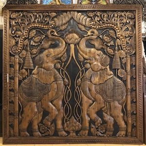 Puede incluir: Un gran panel de madera tallada con dos elefantes enfrentados. Los elefantes est&aacute;n rodeados de dise&ntilde;os florales y foliares detallados, con un fondo oscuro. El marco es de color marr&oacute;n oscuro.
